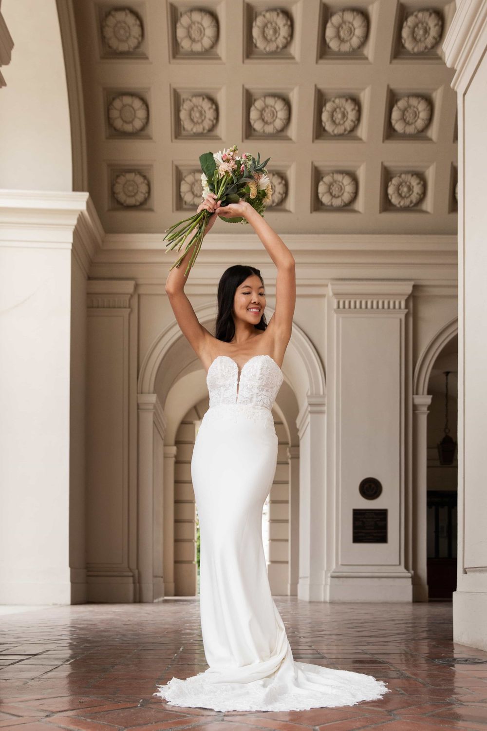 Joyful bride in strapless mermaid wedding gown raises bouquet overhead in ornate arched hall.