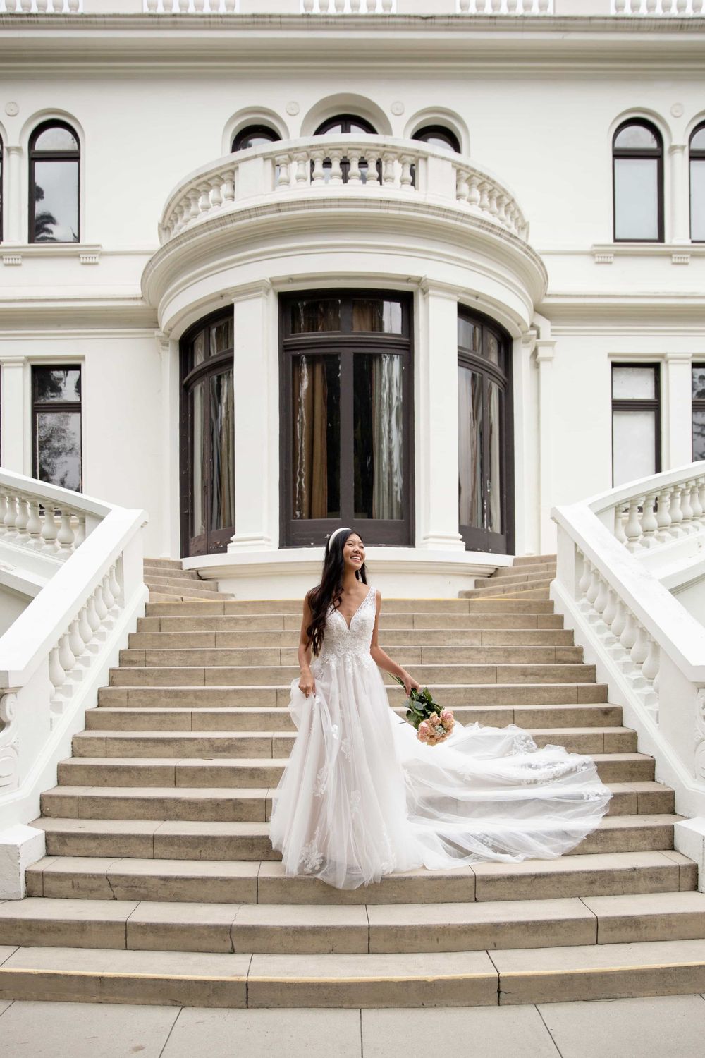Bride in flowing A-line wedding gown descends grand white staircase of elegant mansion.