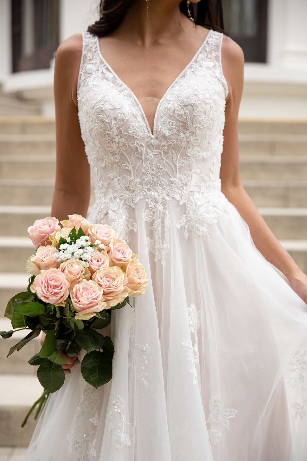 Close-up of a bride in a lace V-neck wedding gown holding a bouquet of soft pink roses.