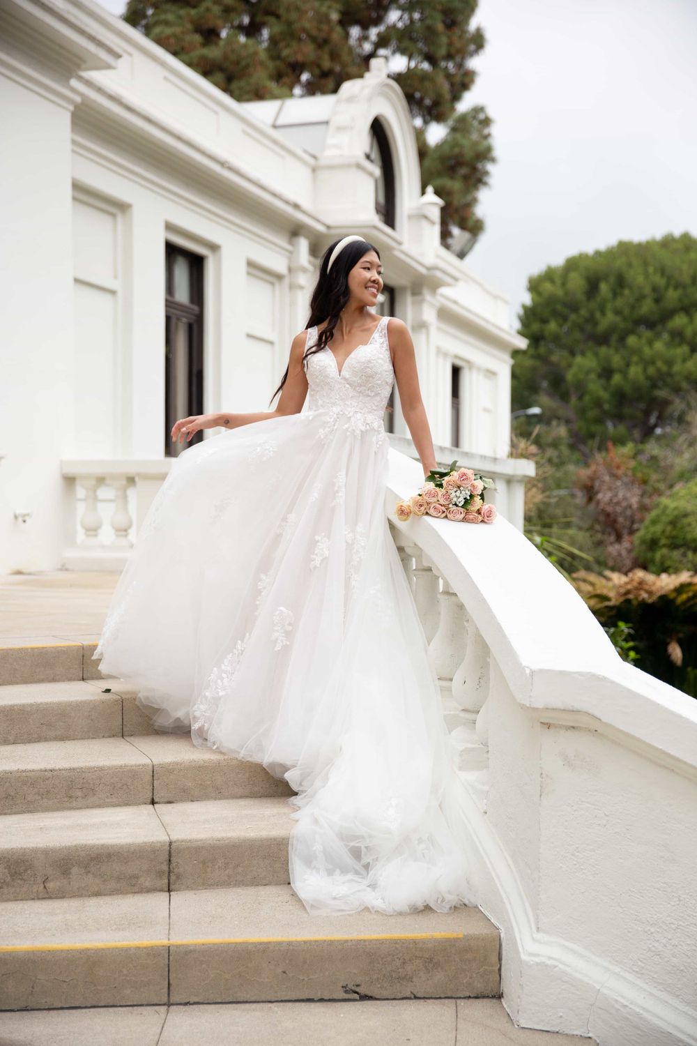 Smiling bride in a lace ballgown wedding dress descending white stone steps holding a pink rose bouquet.