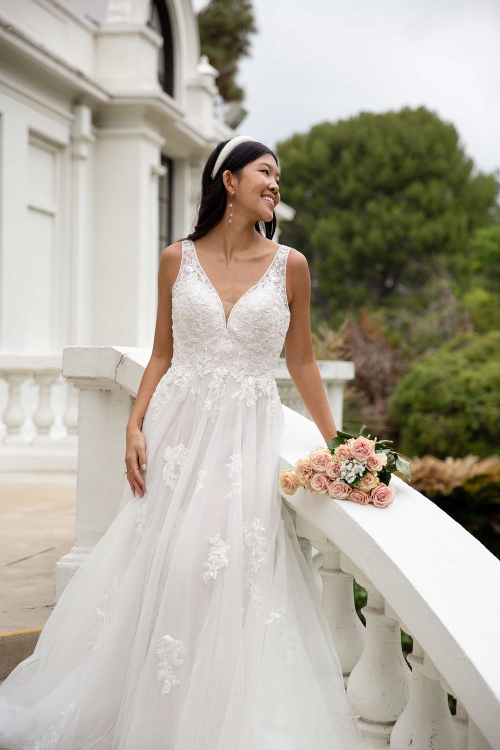 Smiling bride in a lace A-line wedding dress holding pink roses on an elegant white balcony.