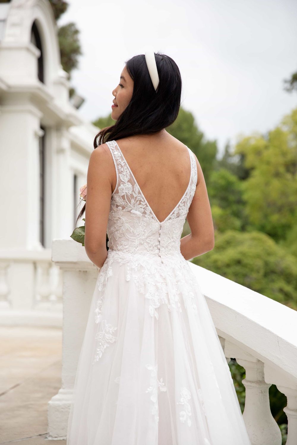 Back view of a bride in a lace V-back wedding dress standing on a white balcony.
