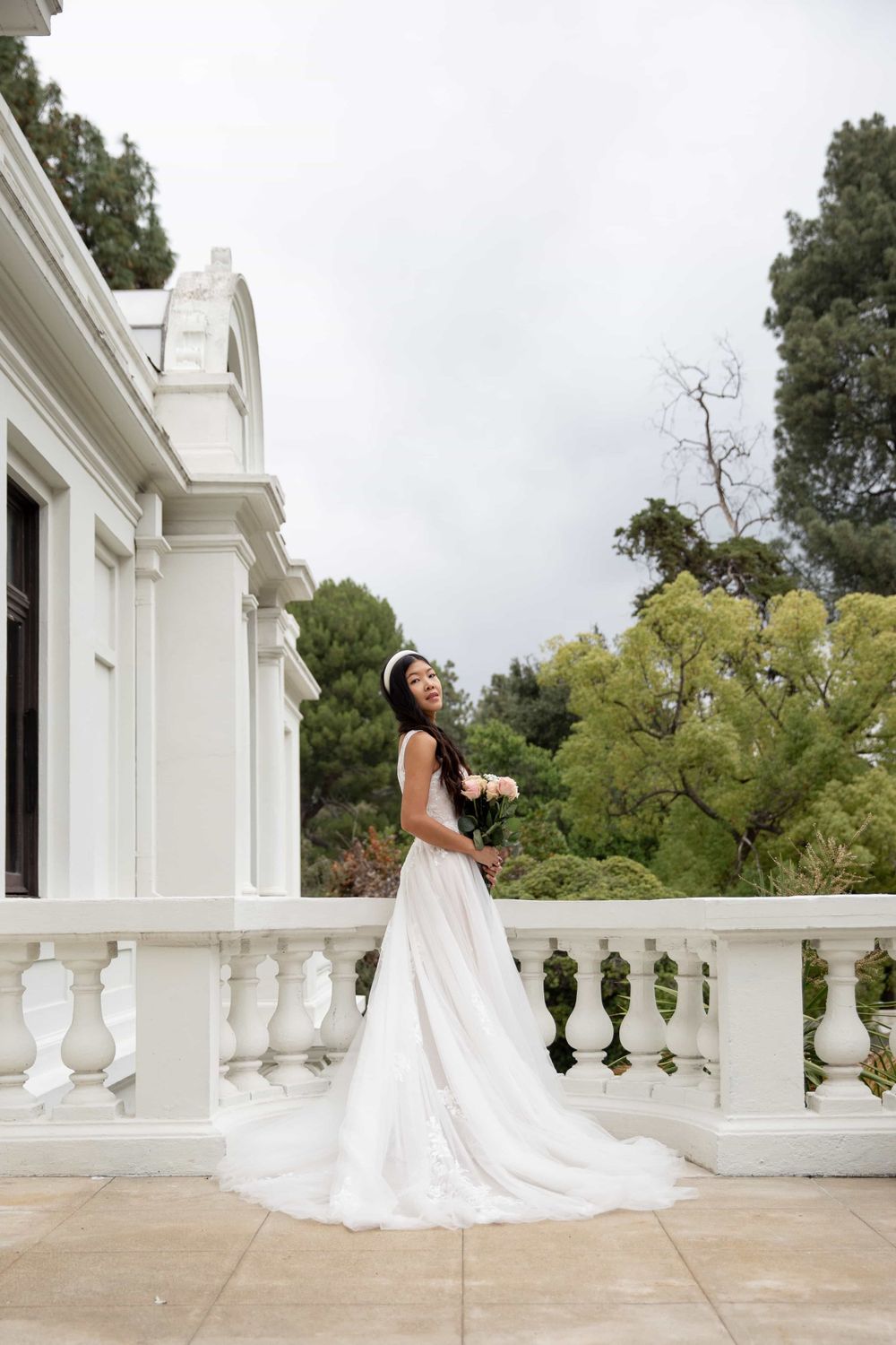 Bride in a flowing white lace wedding gown standing on a grand white balcony surrounded by trees.