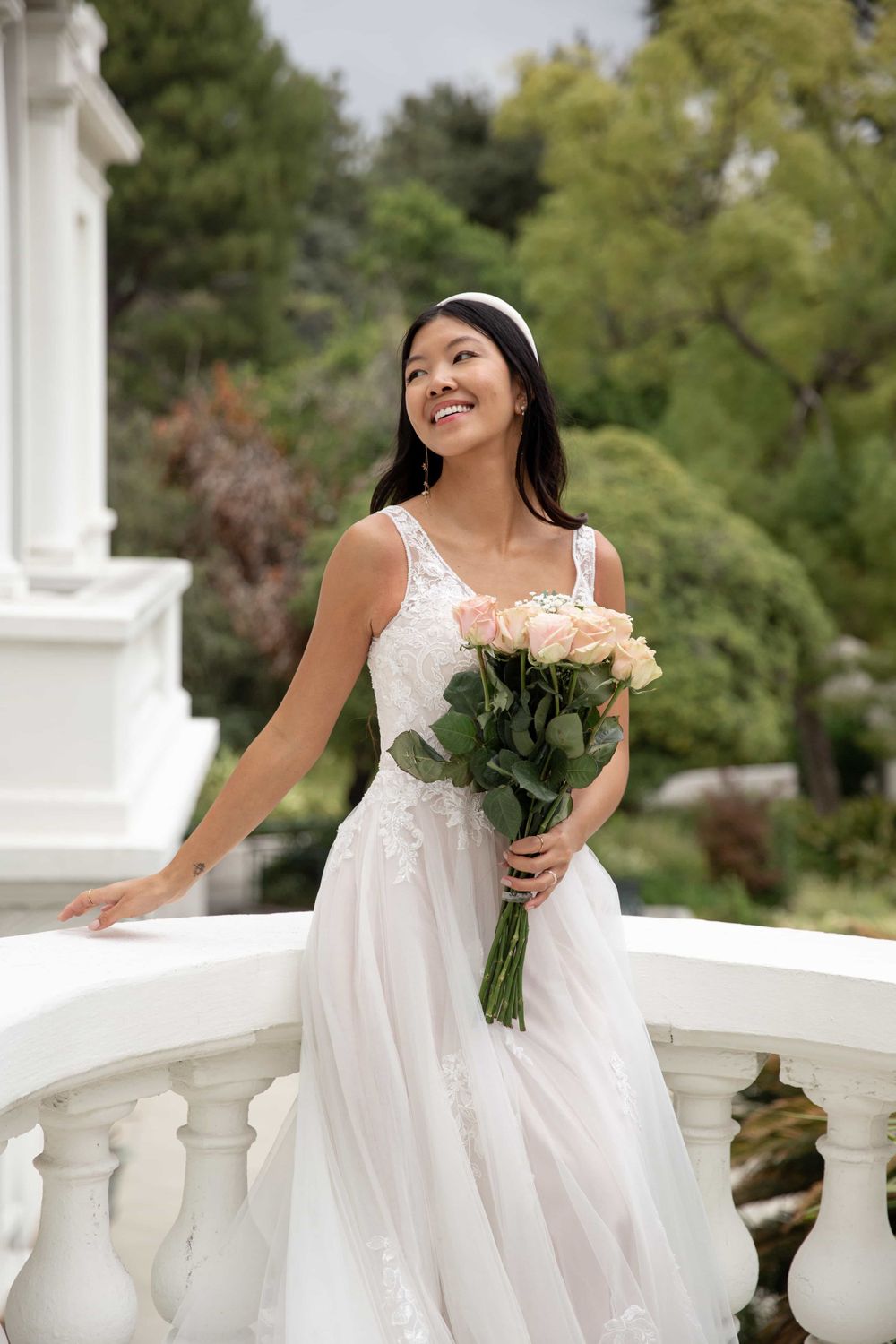 Joyful bride in a lace wedding dress holding pink rose bouquet while standing at a white balcony.