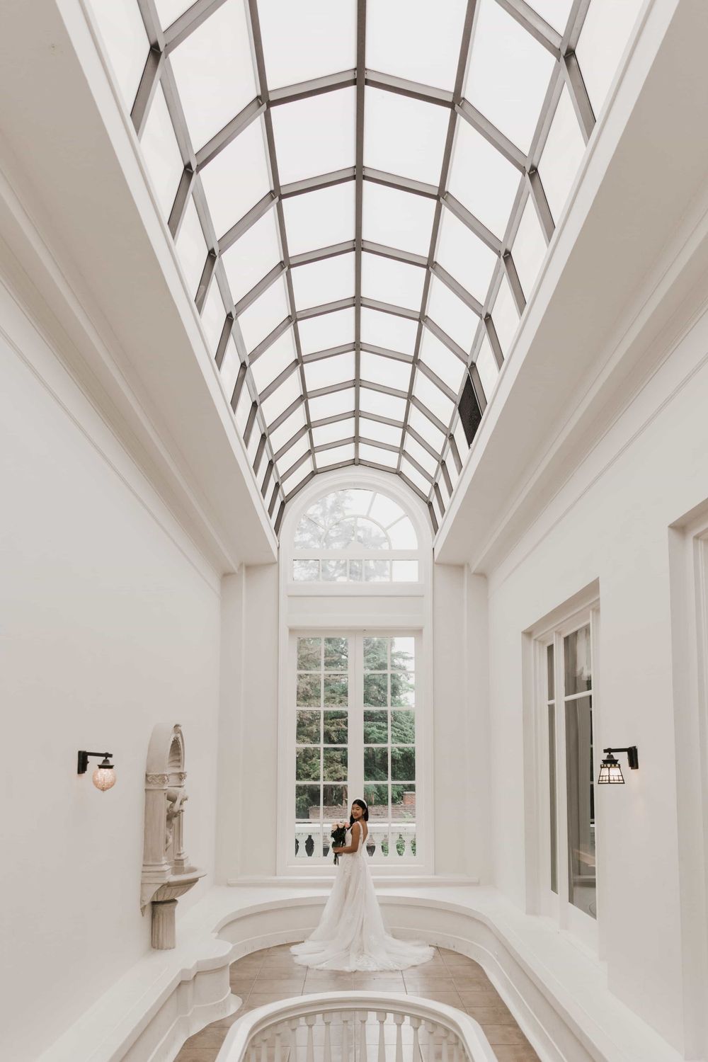 Bride in a white lace gown standing beneath a stunning arched glass ceiling in a grand hall.