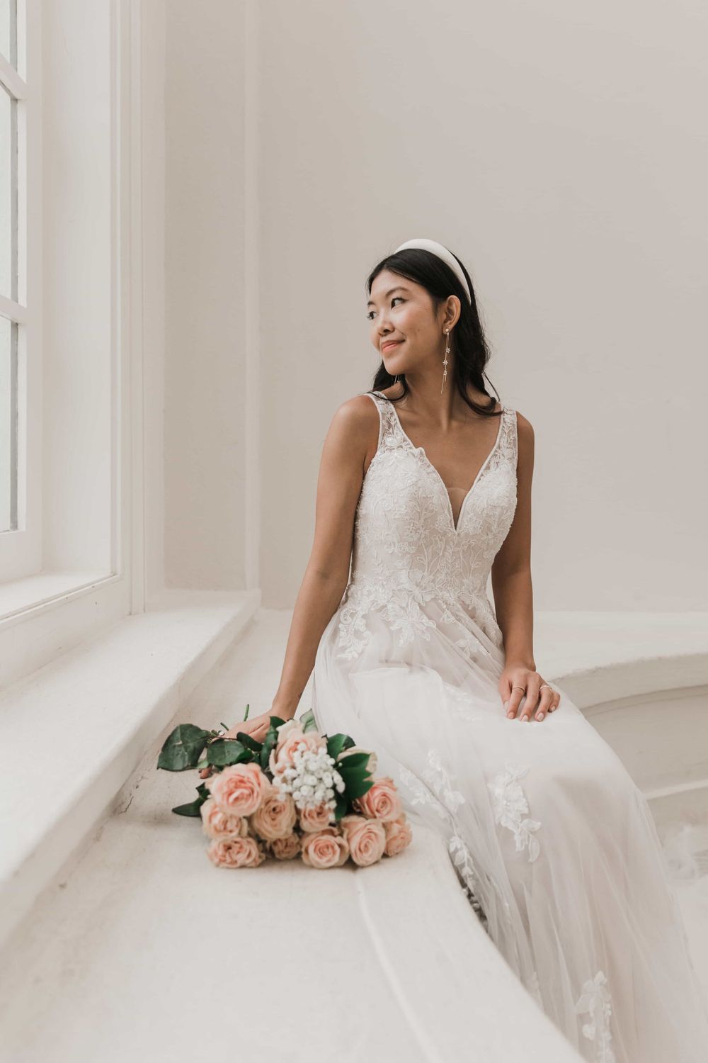 Elegant bride in a lace V-neck wedding dress seated by a window with pink roses beside her.