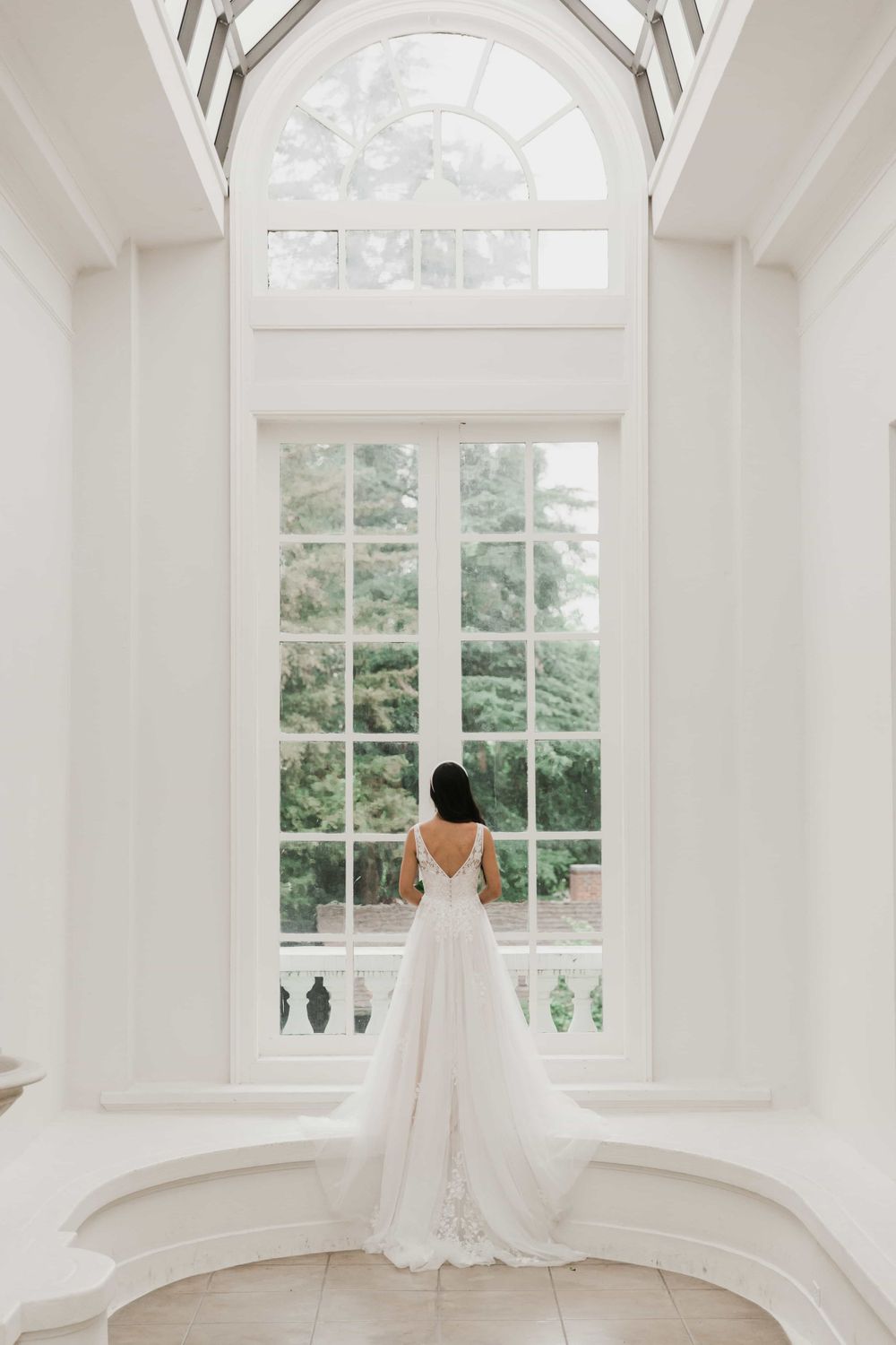 Bride in a lace V-back wedding dress standing before large arched windows in a bright white room.