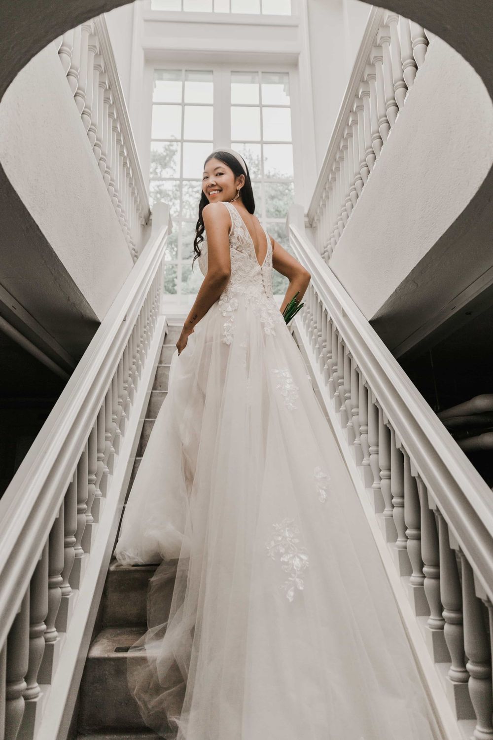 Smiling bride in a lace ballgown looking back while ascending a grand curved staircase.