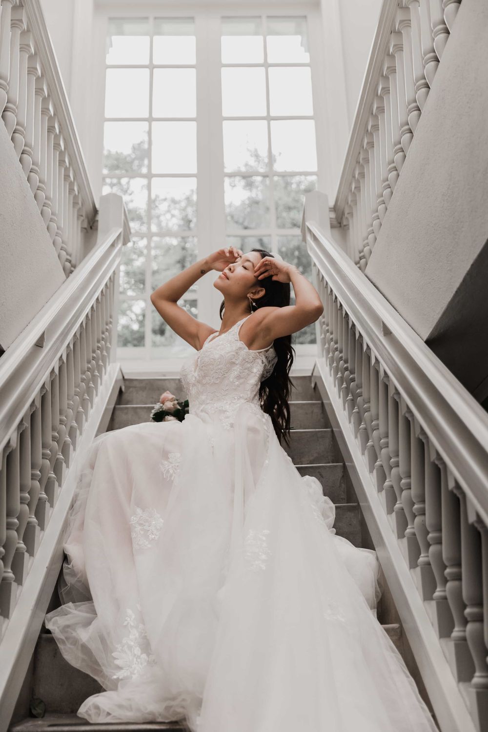 Bride in white lace ball gown sitting on elegant staircase, head tilted back with hands in dark hair.