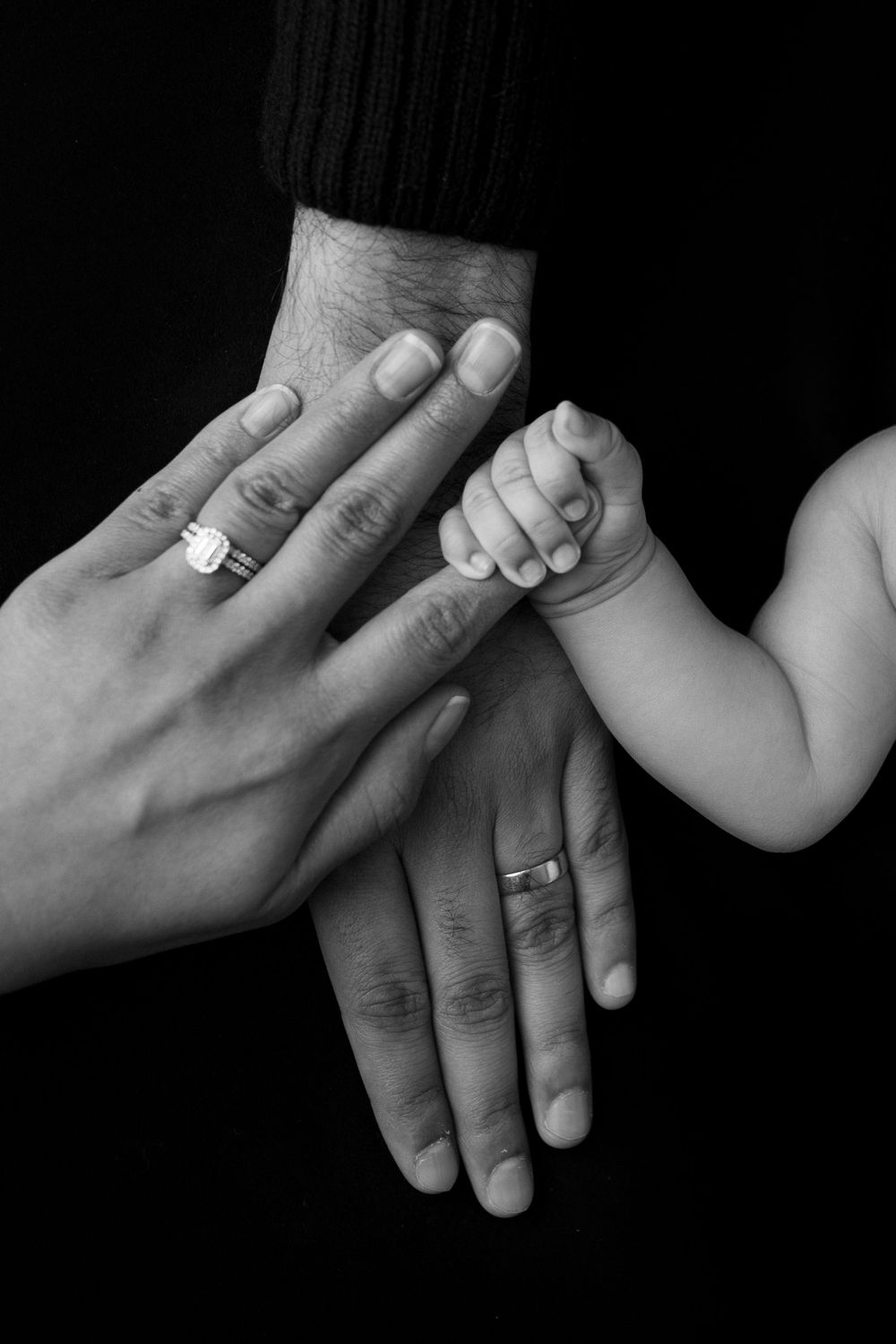Close up of wedding rings and baby hand touching adult hands against black background.