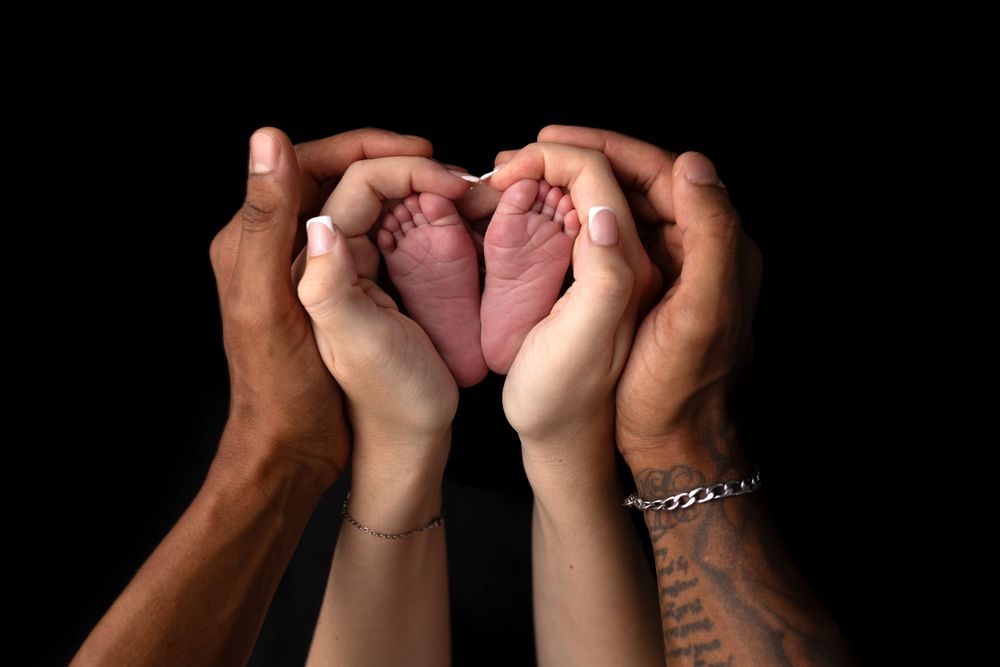 Hands forming heart shape around tiny baby feet against black background.