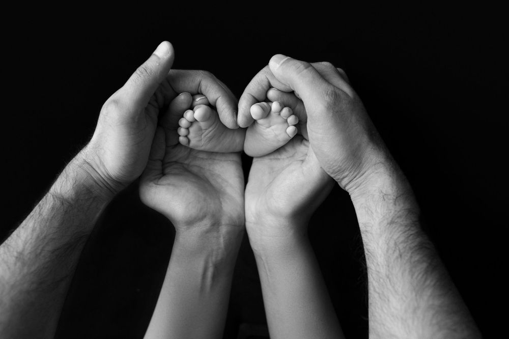 Adult hands holding tiny baby feet forming heart shape against dark background.