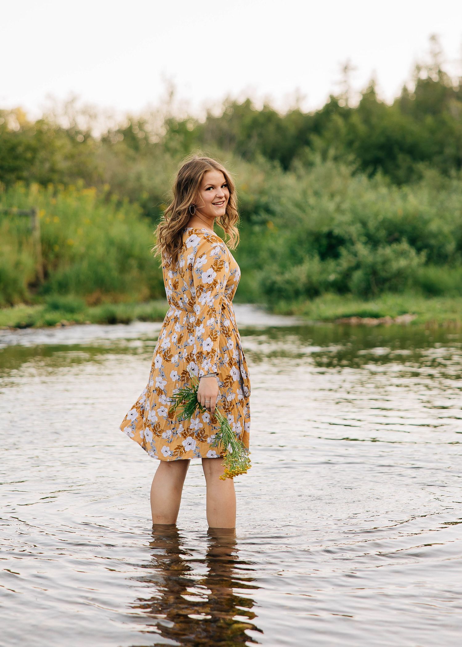 A woman in a floral dress stands in shallow water on a summer day by a serene pond.