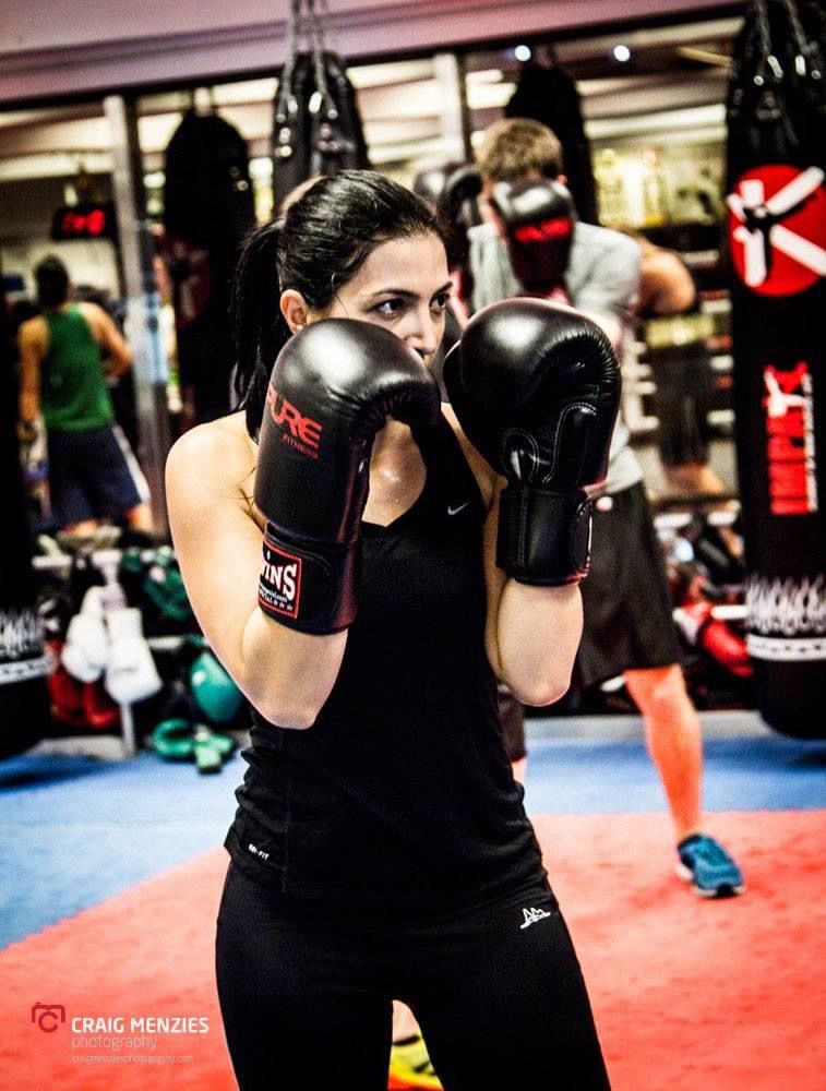 Dark-haired woman in black Nike top practicing boxing stance with Twins gloves in gym.