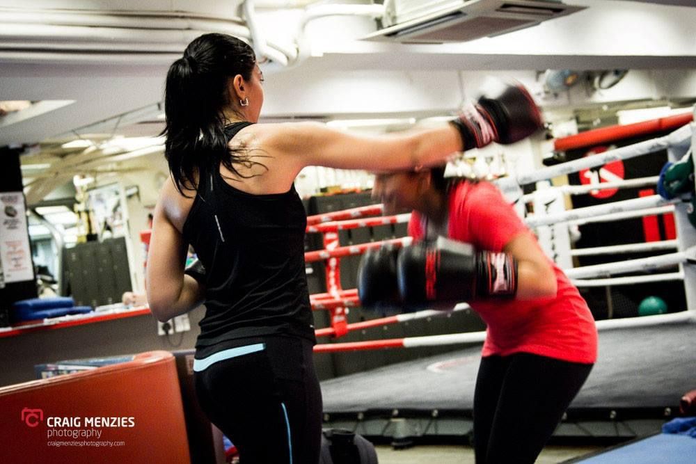 Two women sparring in a boxing gym, one throwing a punch while wearing boxing gloves.