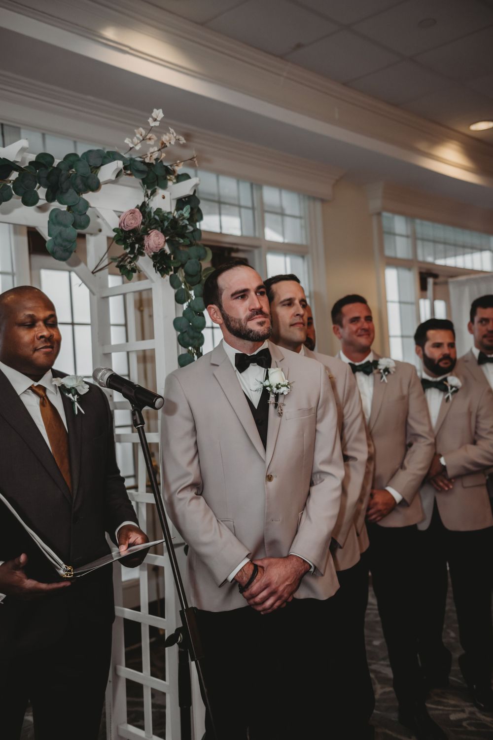Wedding party groomsmen in light suits standing in front of a floral arch during ceremony.
