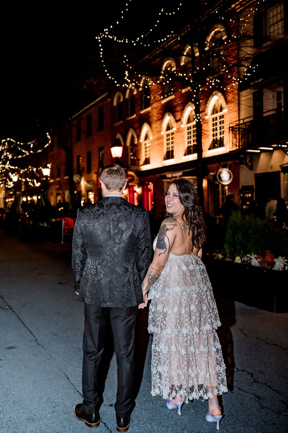 Couple walks along festively lit street at night in formal attire.