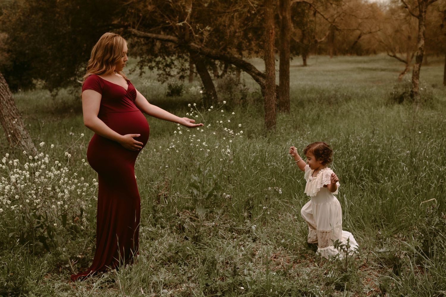 A mother in a burgundy dress and child in white play in a wildflower meadow at sunset.