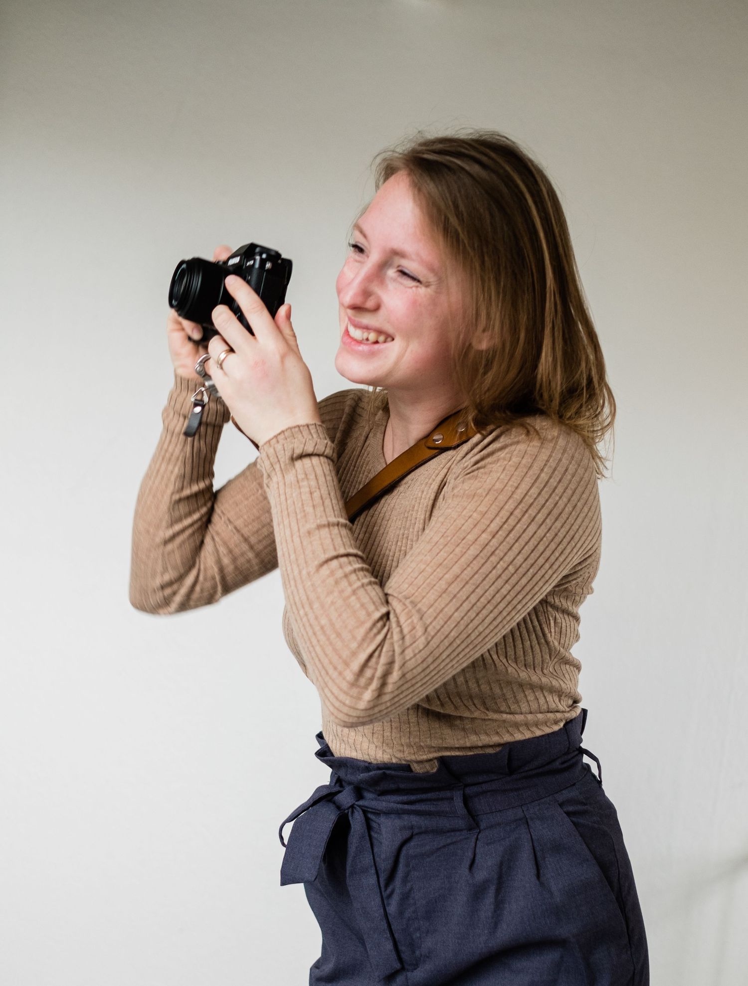 Photographer smiles while holding camera in casual brown sweater and navy pants.