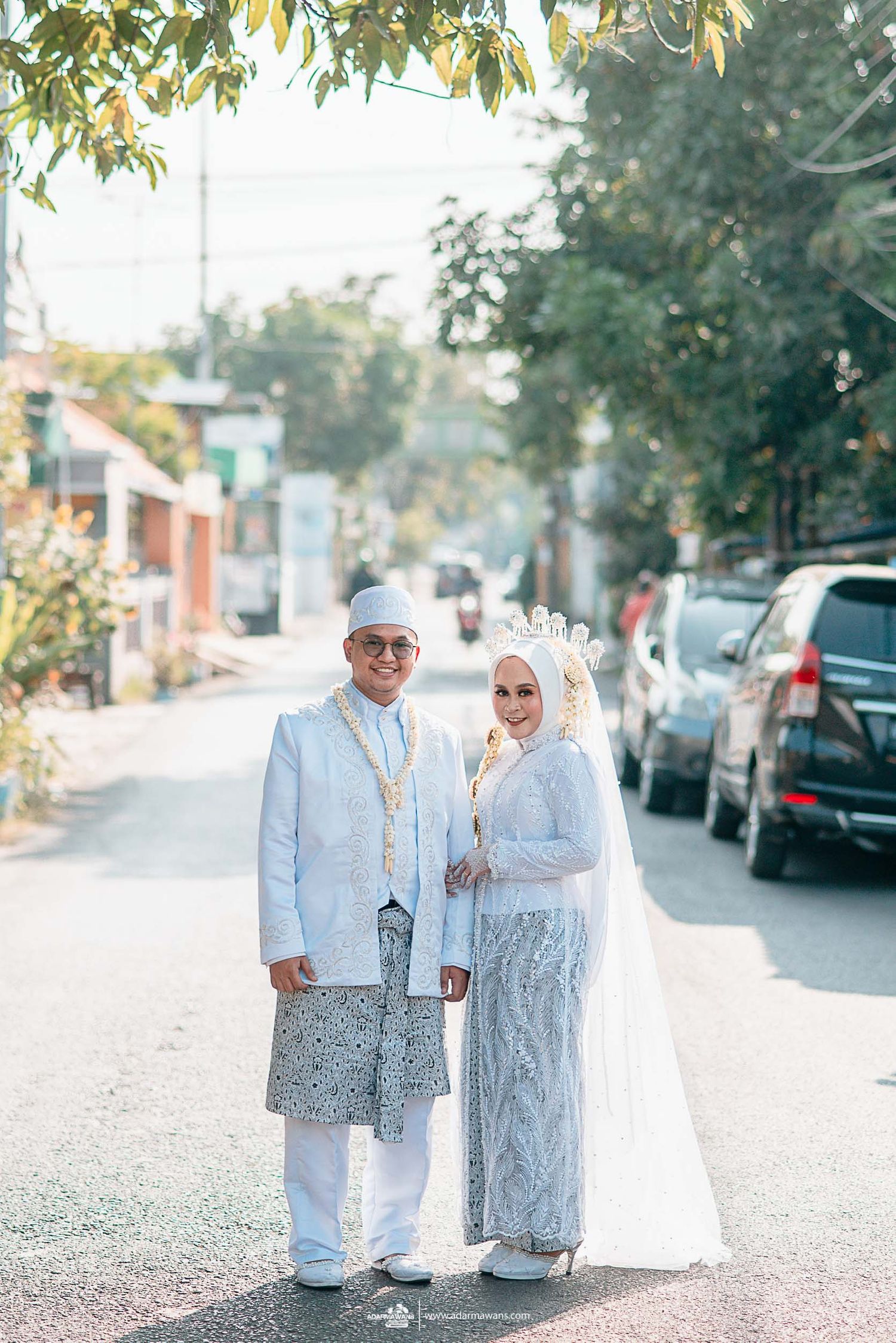 Outdoor wedding portrait on tree-lined street with cars in background.