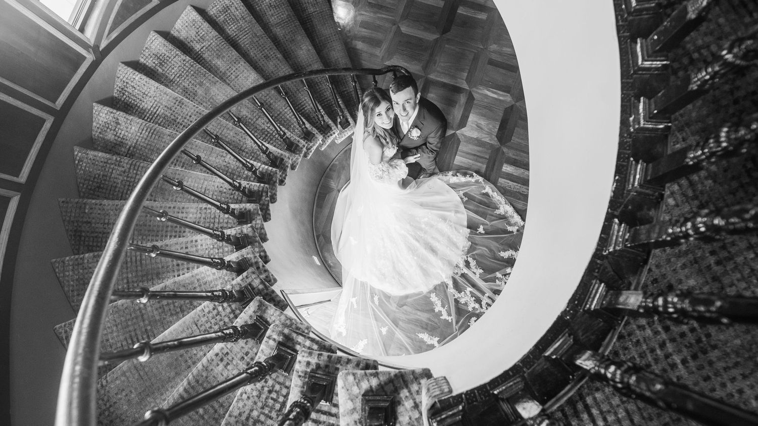 Couple embraced, looking upwards while posing on a spiral staircase.