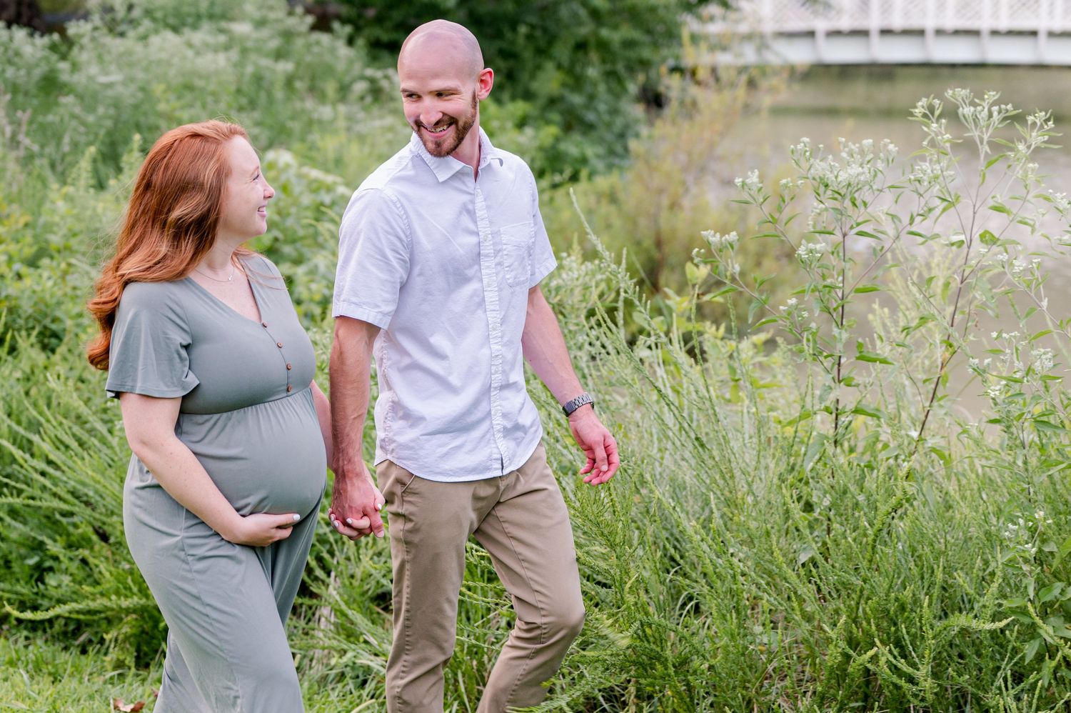 An expecting couple walks together through tall grass on an overcast day in Annapolis.