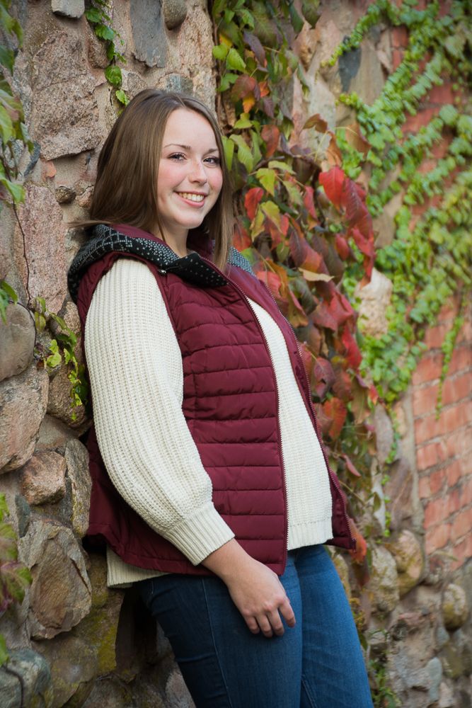 Person in a maroon puffer vest smiles while standing against a stone wall with autumn vines.