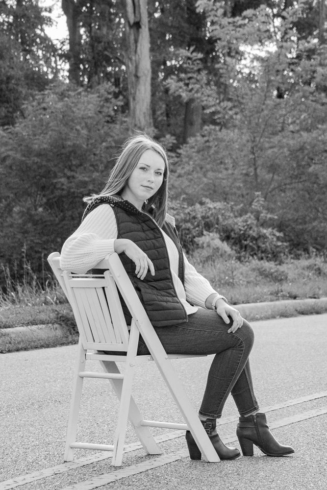 Woman in black vest and white sweater poses on wooden chair in black and white portrait.