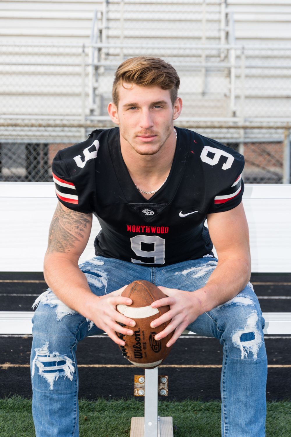 Athletic figure in black jersey holds football while sitting on stadium bleacher.