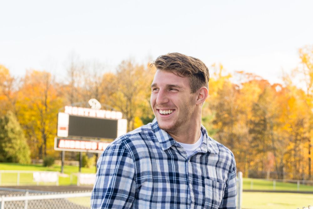 Student athlete in plaid shirt smiles with autumn trees in background.