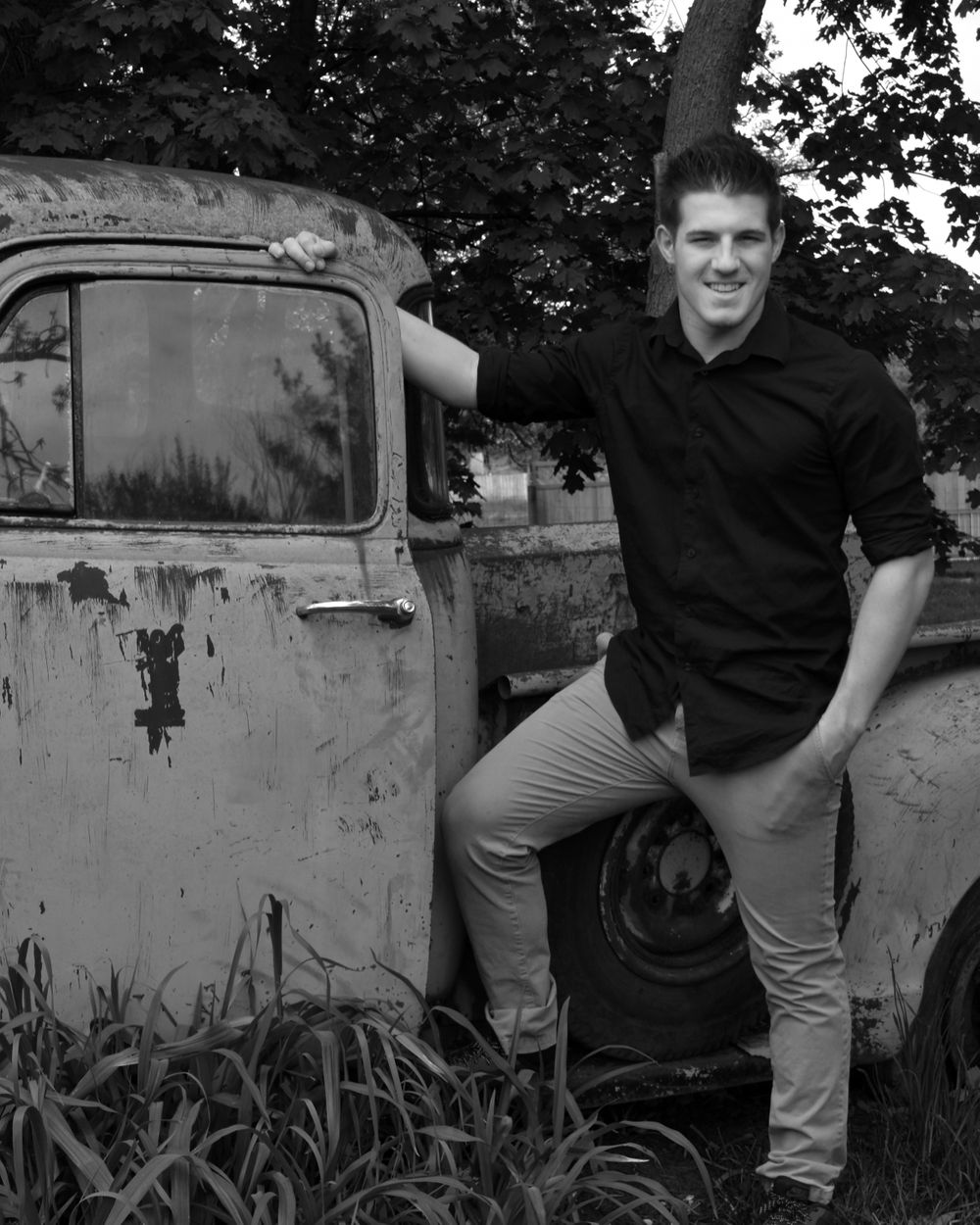 Figure poses in black shirt and khakis next to vintage rusty truck in black and white photo.