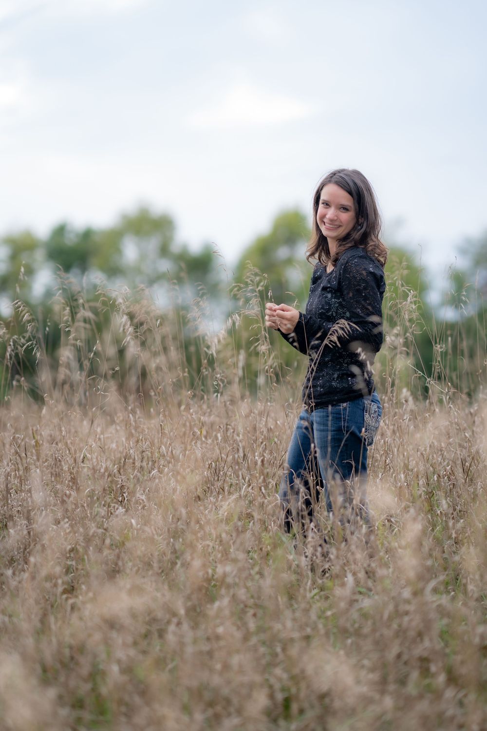 Woman stands among tall dried grasses in blue jeans and black top.