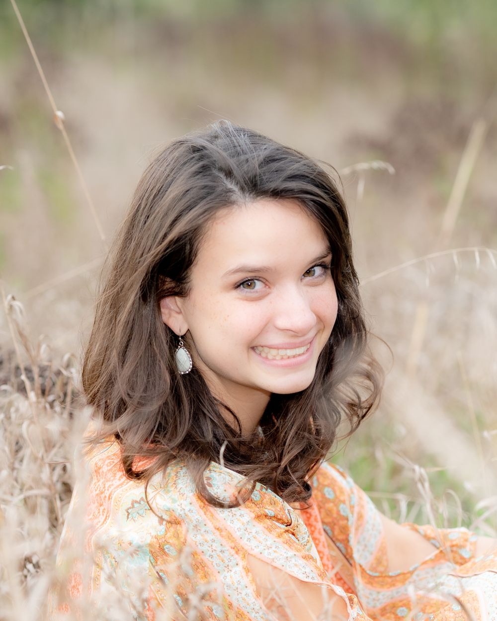 Portrait of woman in orange floral dress sitting in tall grass with natural sunlit background.