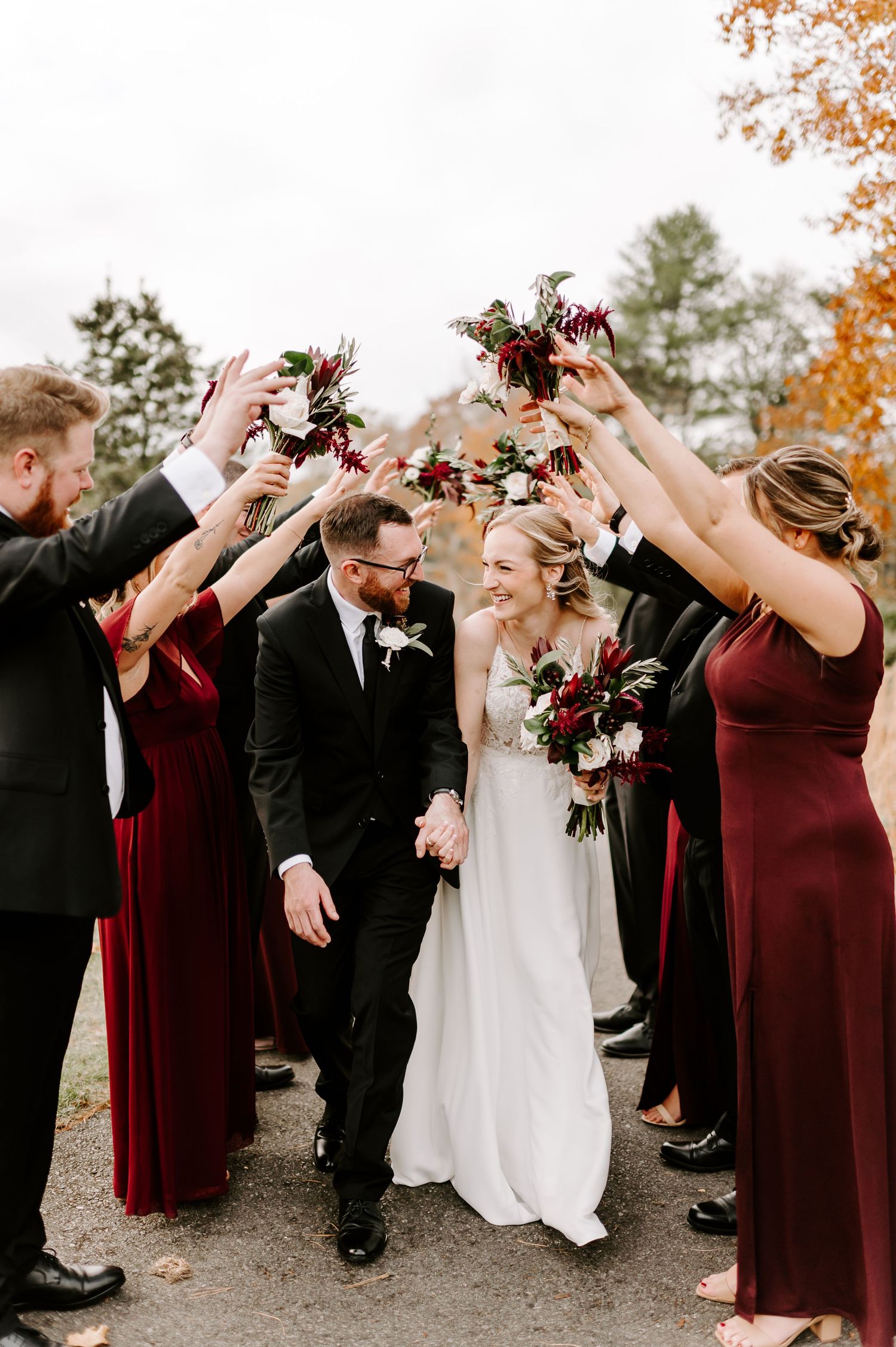 Wedding party celebrates with raised bouquets forming an arch over couple in burgundy and black attire during fall ceremony.