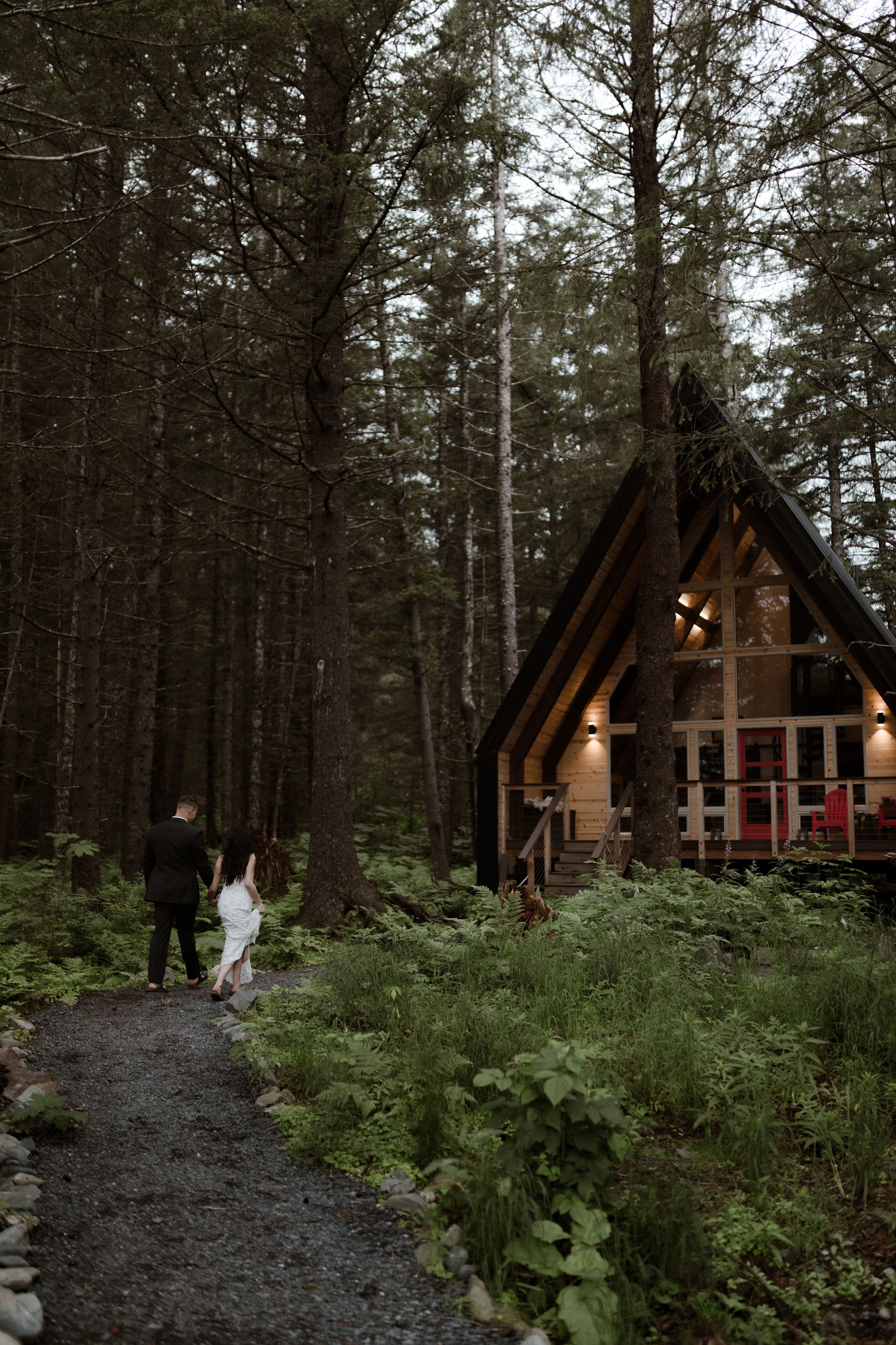 A cozy A-frame cabin glows warmly in a moody forest setting as two figures walk along a stone path at dusk after just getting married.