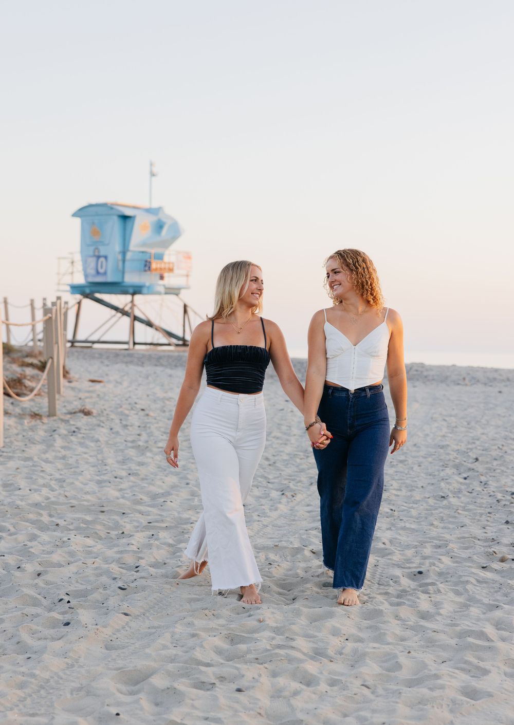 Two women walking and smiling together on beach with lifeguard tower in background.