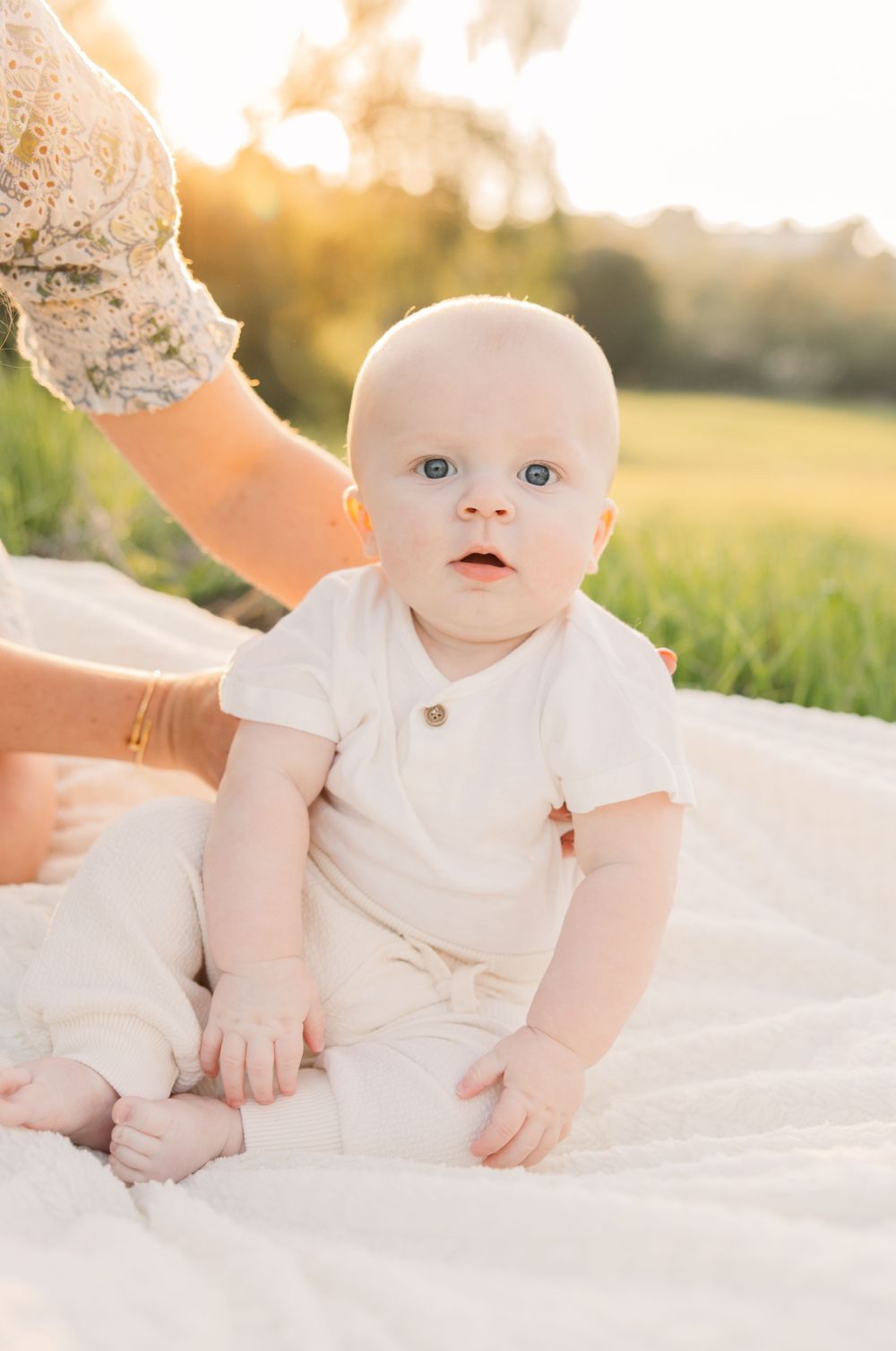 Baby in white onesie sitting on blanket in sunny field during golden hour.