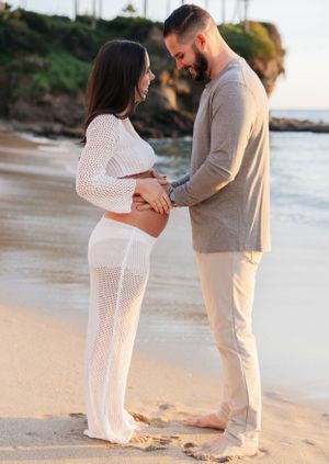 Maternity session on beach with couple in white and beige outfits at sunset.