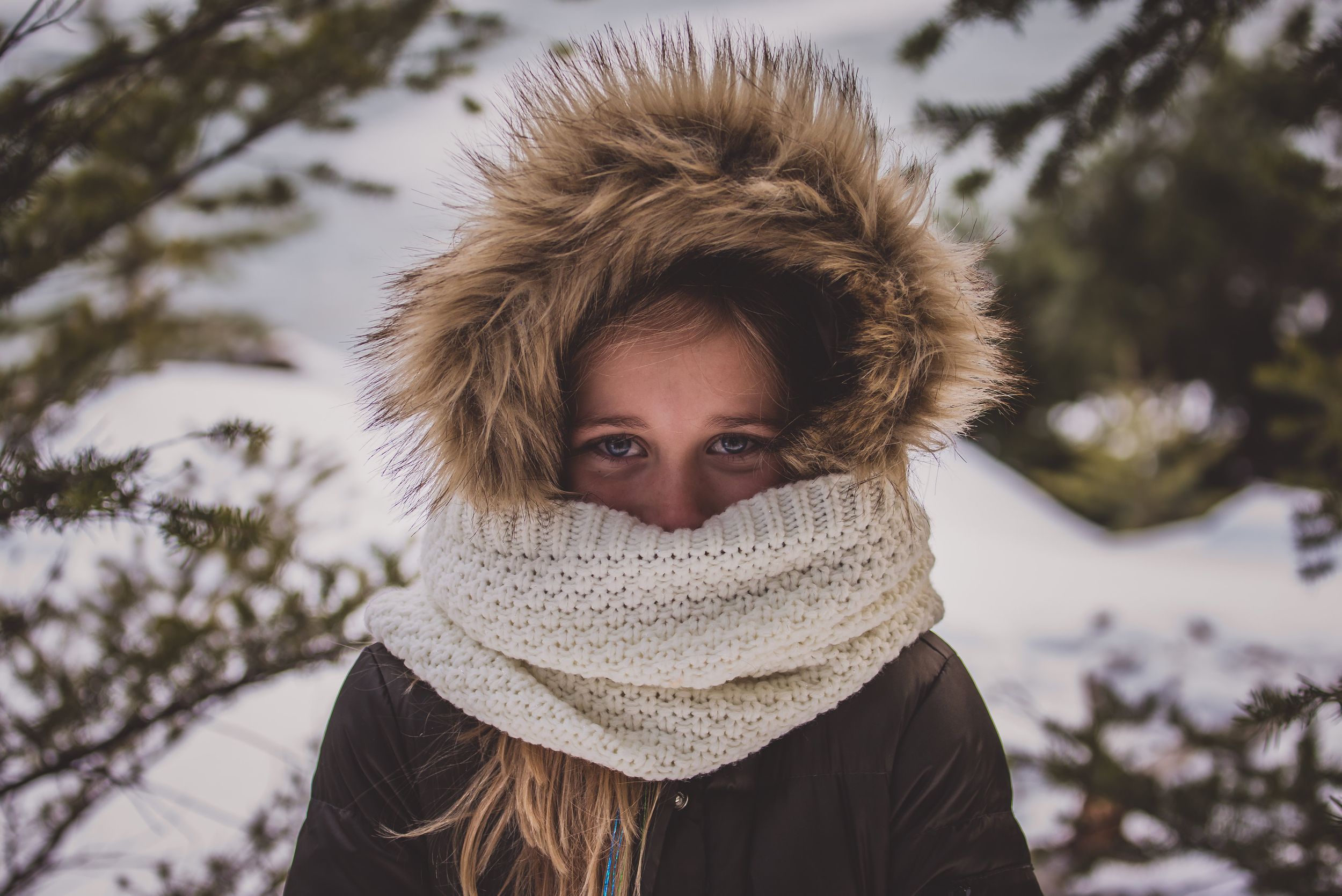 Young woman bundled in fur-trimmed parka and knit scarf standing outdoors in snowy winter conditions.