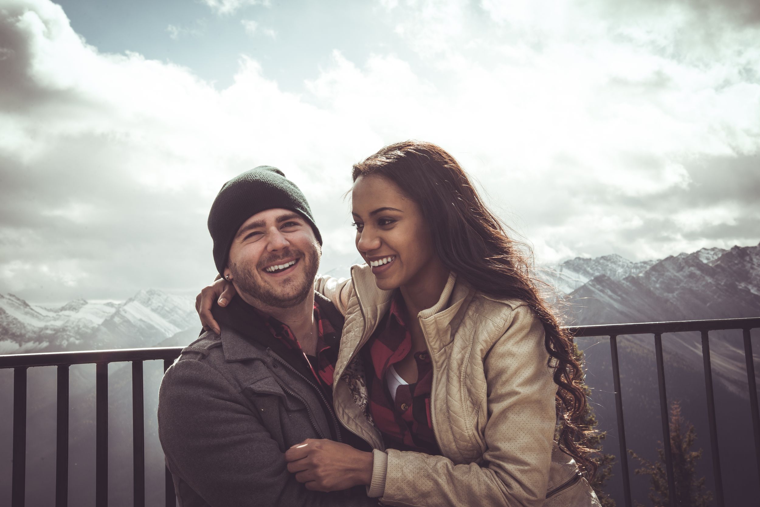 Happy couple embracing and laughing together on a mountain overlook with dramatic cloudy skies and snowy peaks behind them.