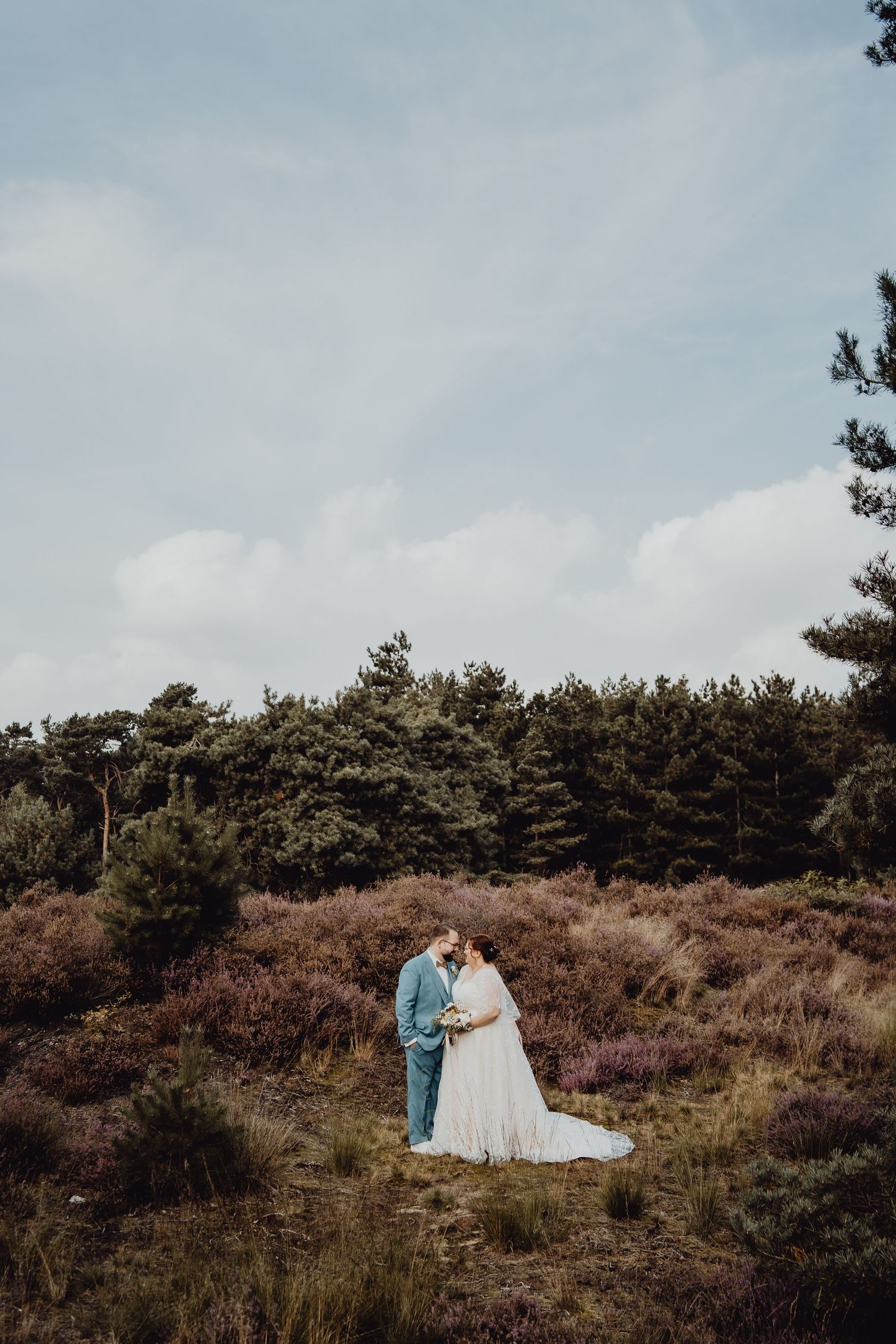 Couple in wedding attire embrace on hillside surrounded by pine trees and purple heather.