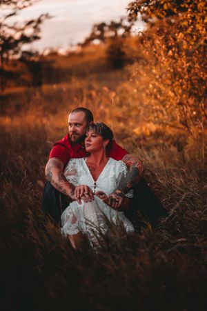 Couple with tattoos sitting in golden tall grass field embracing during a warm sunset.