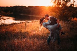 Couple sharing romantic dip kiss in golden sunlit meadow near lake.