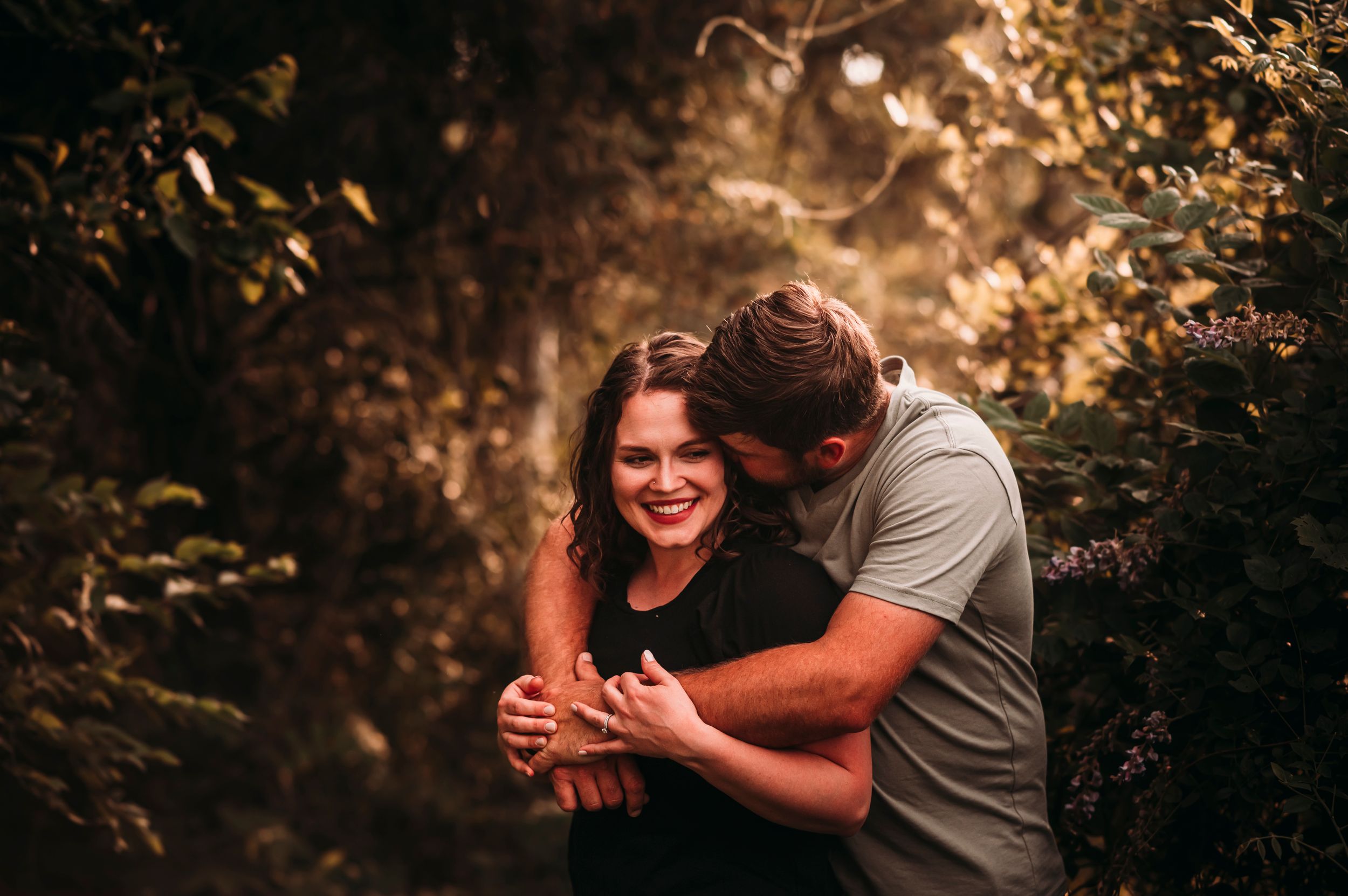 Happy couple embracing and laughing together surrounded by lush green foliage in warm golden sunlight.