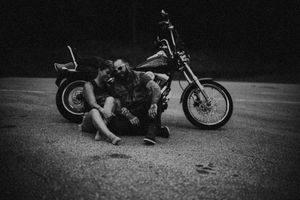 Couple sitting together on pavement leaning against a motorcycle in a black and white night photo.