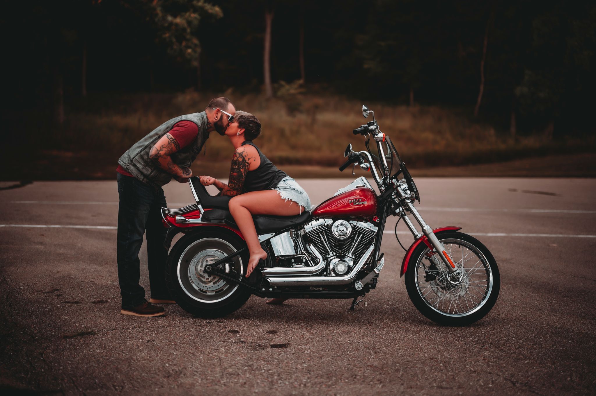 A couple shares a romantic kiss beside a red Harley-Davidson motorcycle in a parking lot surrounded by trees.