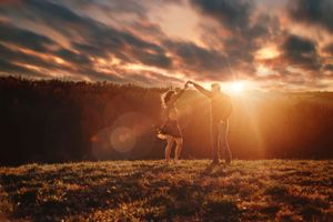 Couple dancing together in a golden sunset field with dramatic cloudy sky backdrop.
