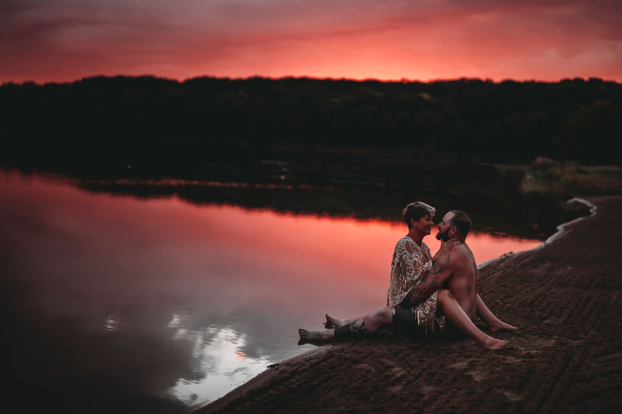 Romantic couple sitting by a reflective lake sharing a kiss under a dramatic red sunset sky.