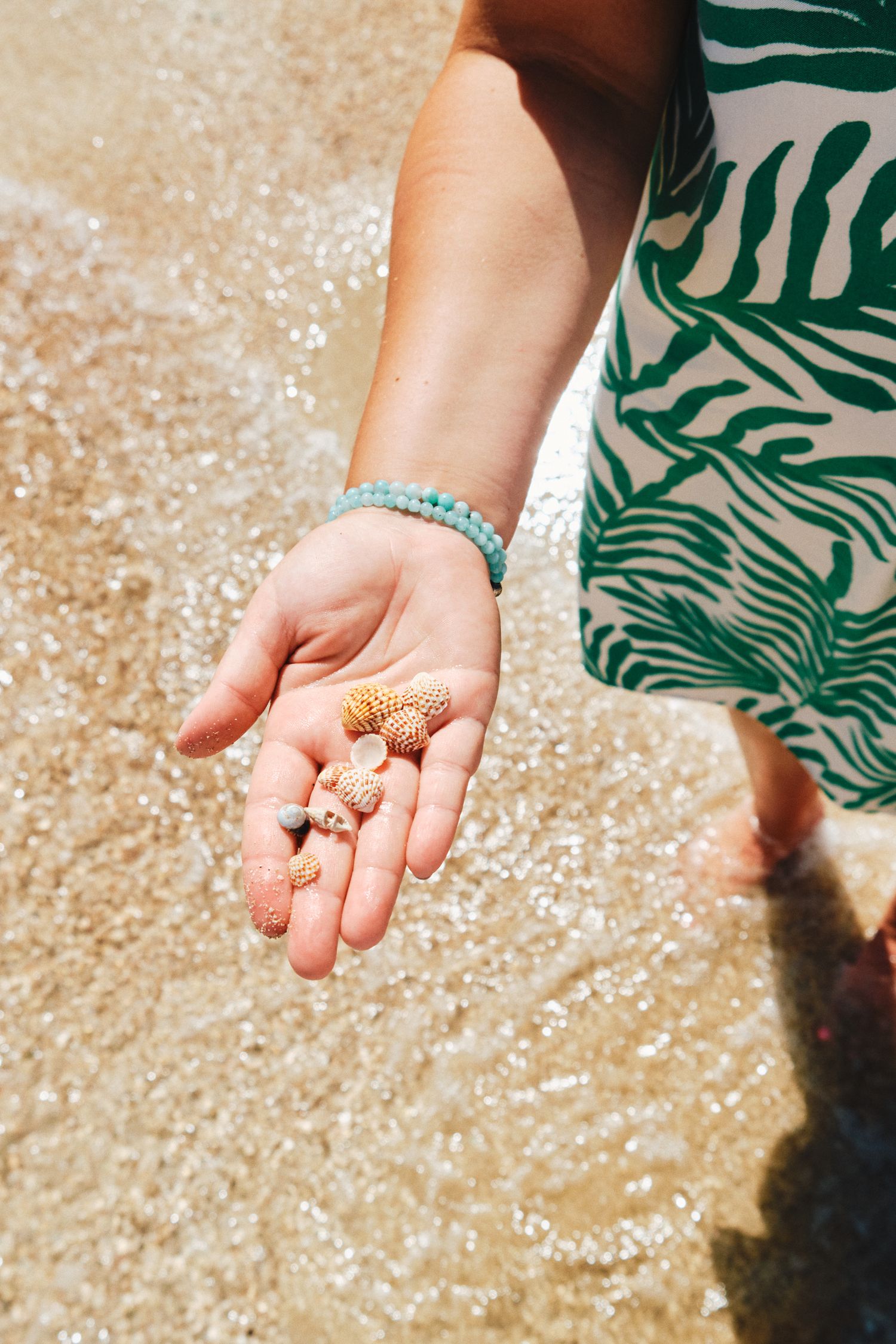 Open palm holding tiny seashells while wearing tropical print dress on sandy beach.