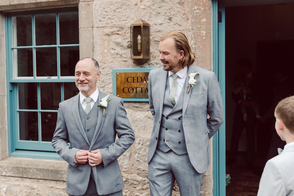 Groomsmen in matching grey suits gather outside a rustic stone building.
